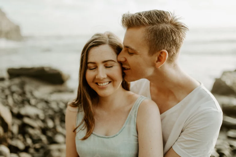 couple cuddling and kissing each other as they get engagement photos on the beach in Auckland